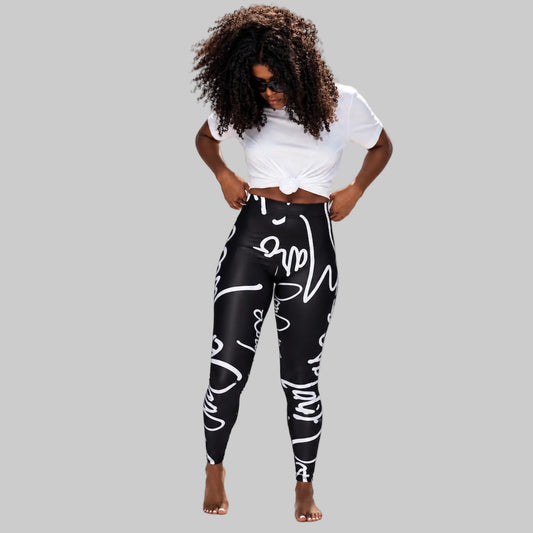A woman modeling high-waisted black leggings with a white abstract graffiti-style print, paired with a white cropped tie-front t-shirt, against a light gray studio background.