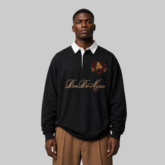 A man wearing a black long-sleeve polo sweatshirt with a white collar, gold 'Don DeMarco' script on the chest, and a red/gold crest, paired with brown trousers, against a neutral gray background.